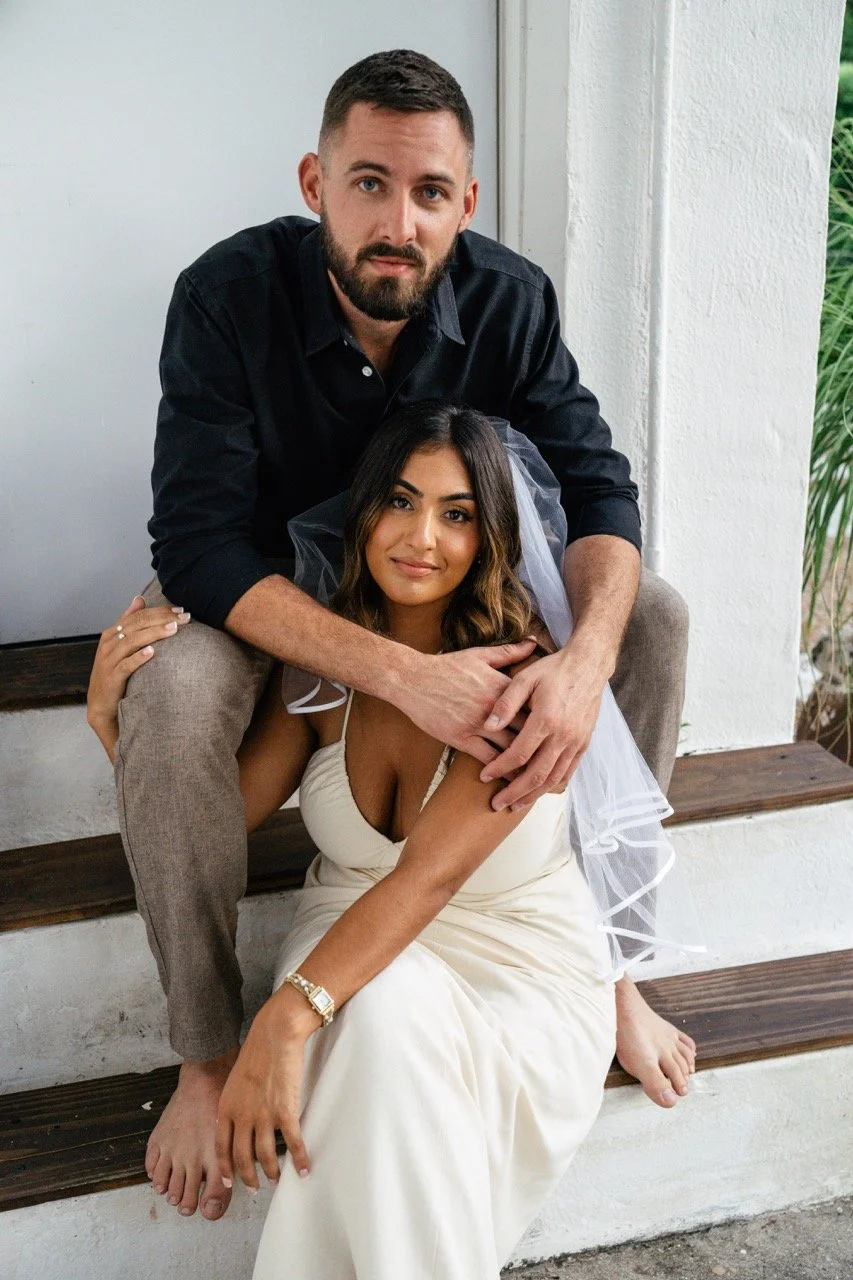 A man and a woman sitting on wooden stairs, with the man sitting behind and hugging the woman. The woman is wearing a white dress and a veil, indicating it may be a wedding. The man is dressed in a black shirt and gray pants. They are outdoors near a