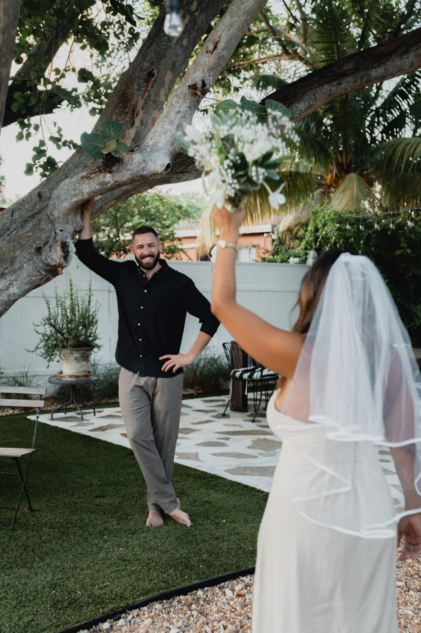 A bride in a white dress and veil cheerfully tosses a bouquet to a smiling man in a backyard. The man is wearing a black shirt and khaki pants, standing barefoot on the grass under a large tree.