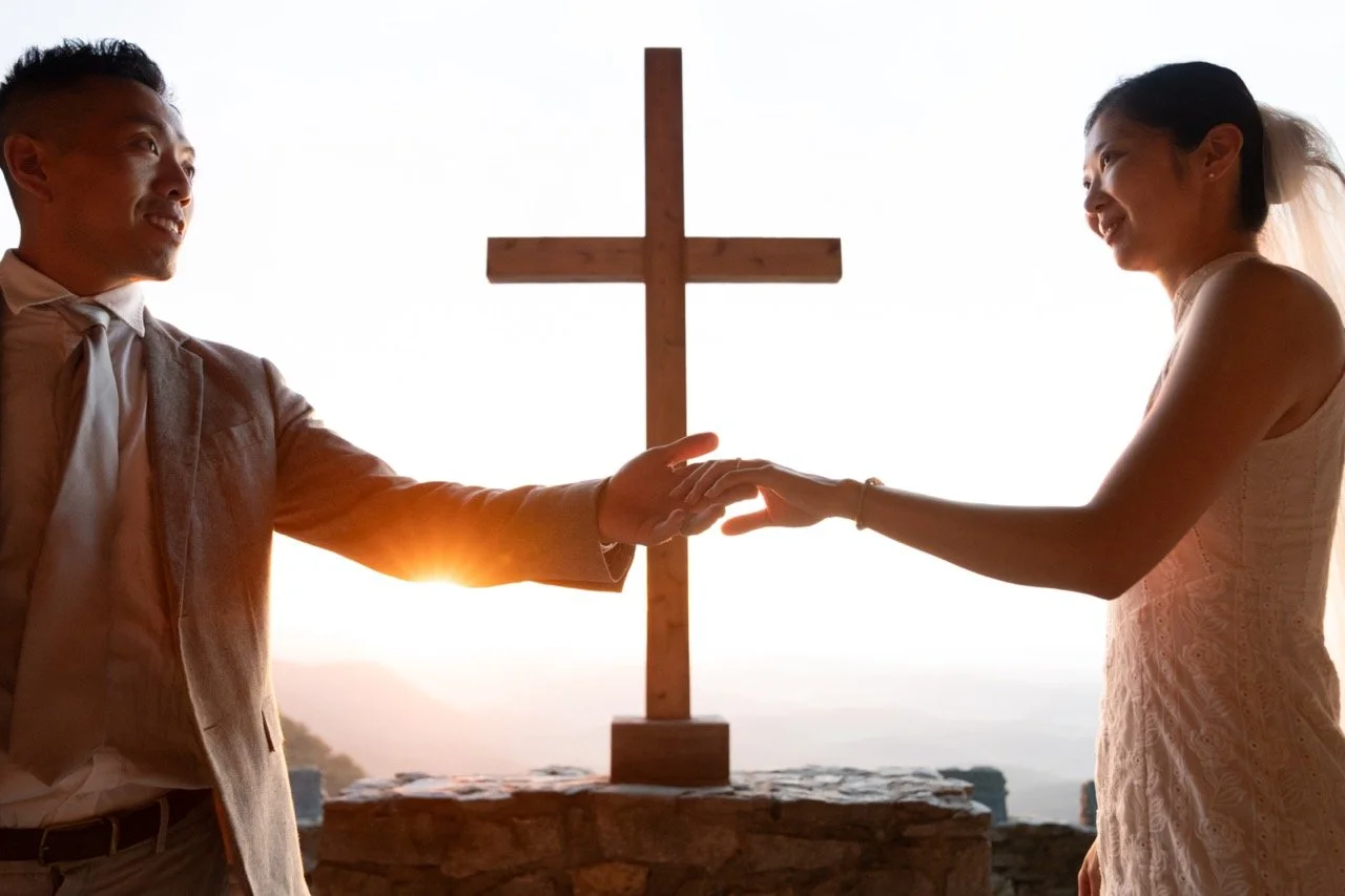 A couple in wedding attire holding hands in front of a wooden cross during sunset.