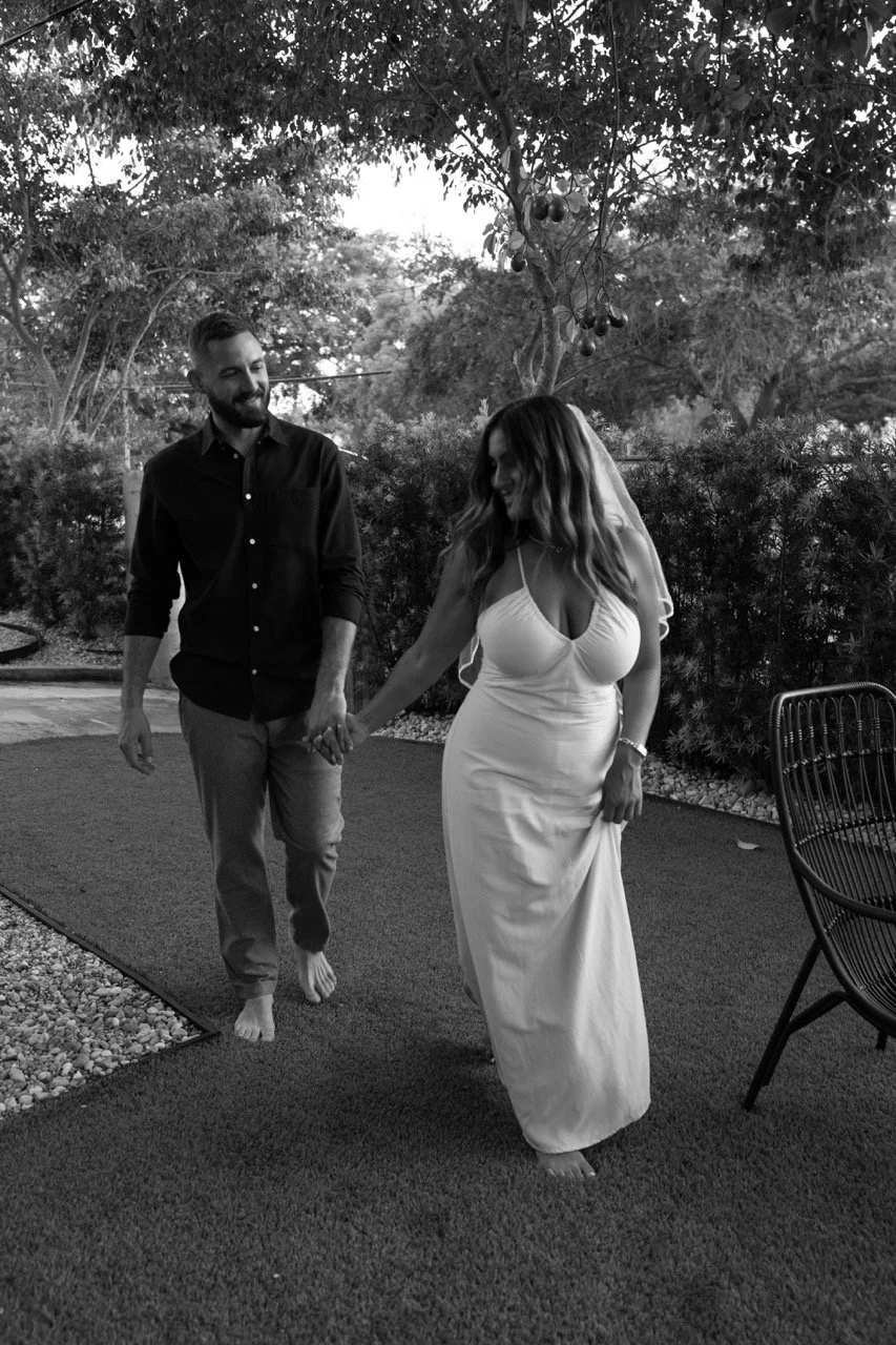 A couple holding hands and walking barefoot outdoors, smiling, with trees and garden in the background, in black and white.