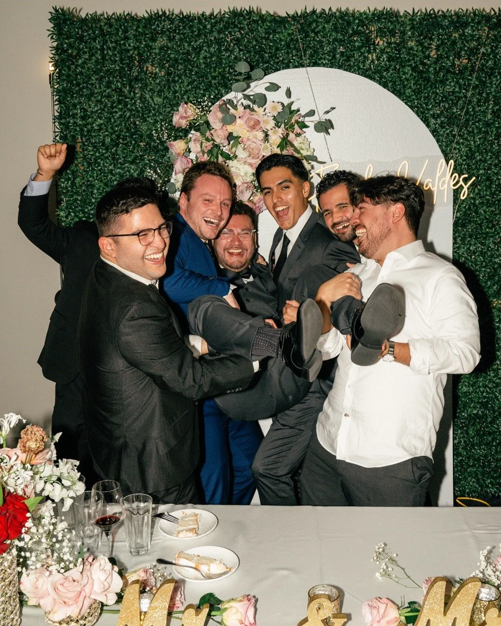 A group of six men celebrating at a wedding reception, lifting one person in the air and smiling. There is a decorated backdrop with flowers and a circular sign in the background.