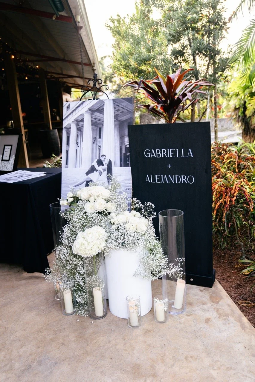 Outdoor wedding display with blackboard sign reading 'Gabriella + Alejandro', a black-and-white photo of a couple, white floral arrangements, white candles in glass holders, and lush greenery in the background.