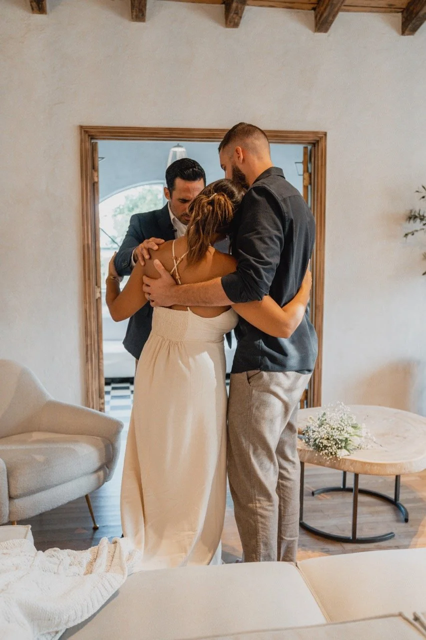 Three people embrace in a warm hug inside a cozy, rustic room.
