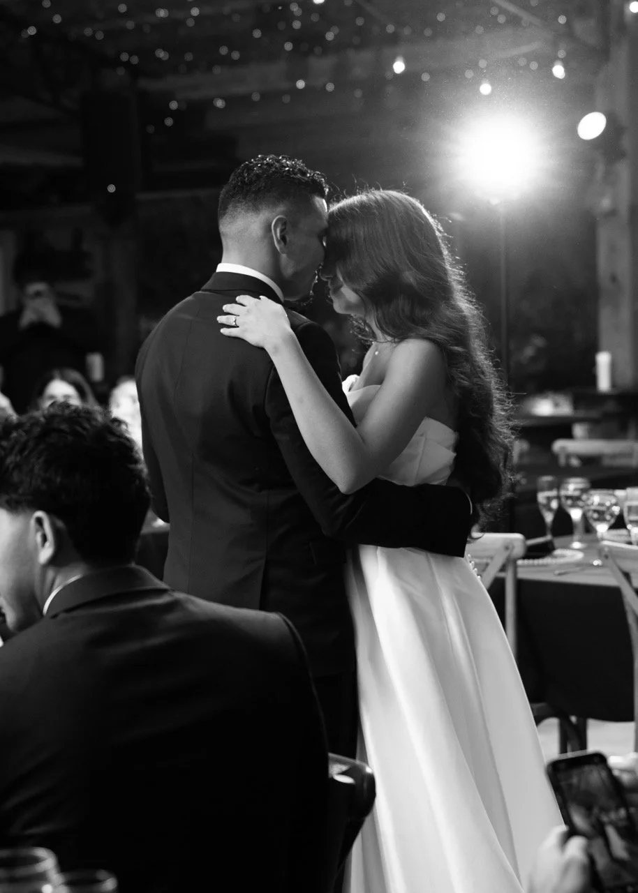 Black and white photo of a couple dancing closely at their wedding reception, with guests and tables in the background.