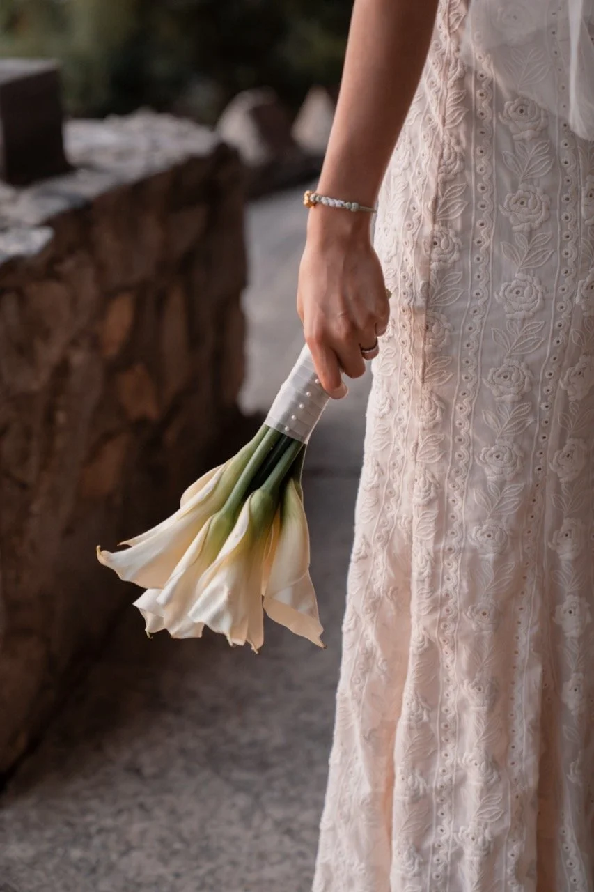 A person dressed in a white, embroidered dress with floral patterns, holding a bouquet of white calla lilies by their side.