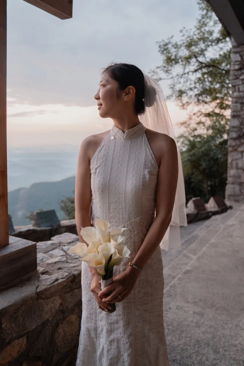 A bride in a white lace wedding dress and veil holding a bouquet of white calla lilies, standing outdoors at sunset with a mountainous landscape in the background.