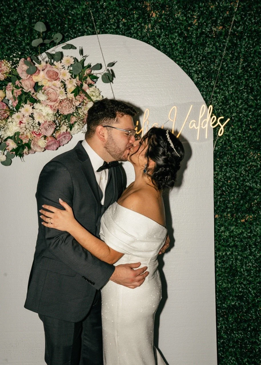 A newlywed couple sharing a kiss at their wedding, standing in front of a floral backdrop and greenery wall with elegant gold lettering.