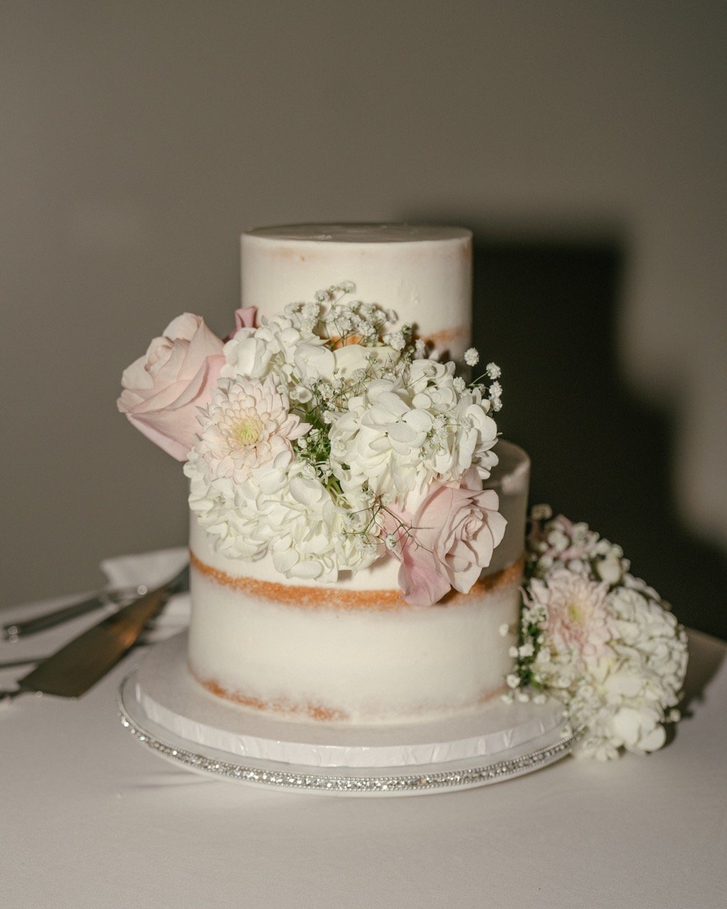 Two-tier wedding cake decorated with fresh white and pale pink flowers, placed on a silver decorative cake stand with a knife and server beside it.