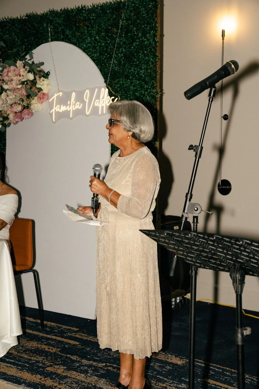 An elderly woman with gray hair, wearing glasses and a beige dress, is standing with a microphone, giving a speech at an event. There is a podium with a music stand nearby, and a decorative backdrop with flowers and a neon sign that reads 'Familia Va