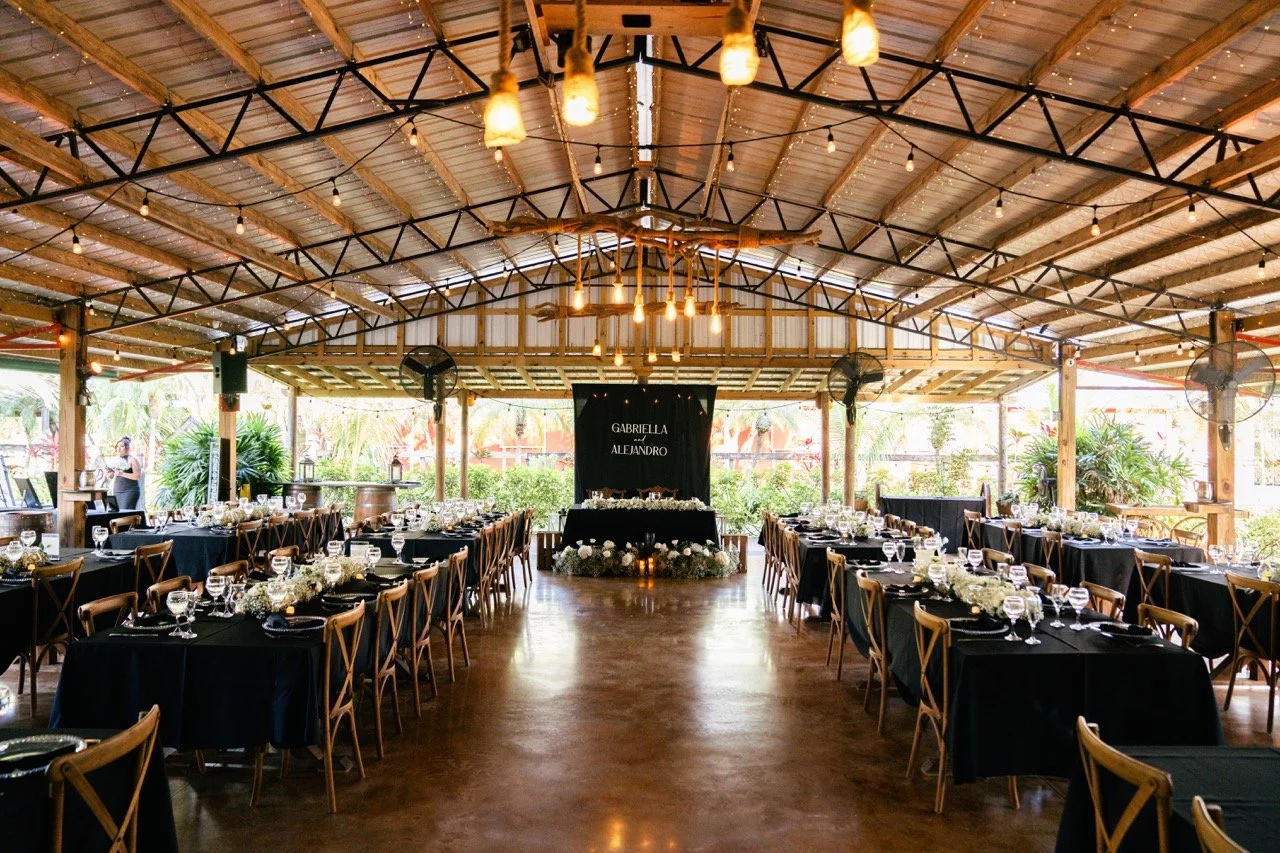 Wedding reception setup in a rustic, open-air venue with wooden beams, string lights, and decorated tables with black tablecloths, glassware, and floral centerpieces, featuring a dance floor and a backdrop with names Gabriella and Alejandro.