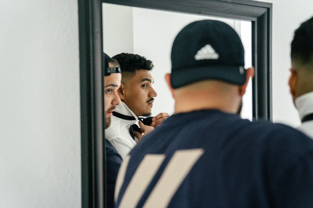 Three young men, one adjusting his hoodie and two wearing caps, are looking at their reflection in a mirror.