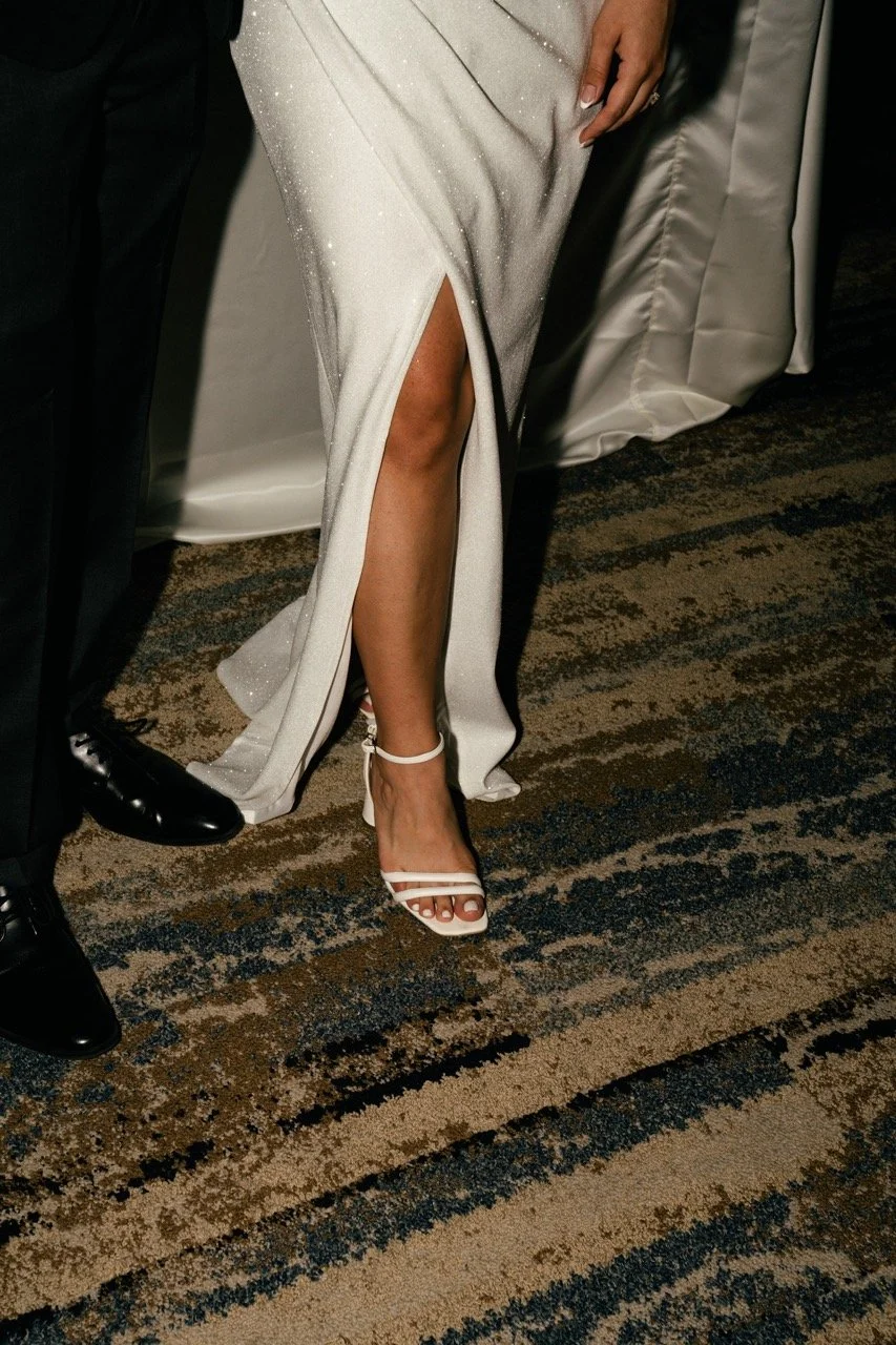 Close-up of a woman's leg in a white, sparkly gown with a high slit, showing her high-heeled white sandals. Part of a man standing next to her, dressed in black, is also visible.