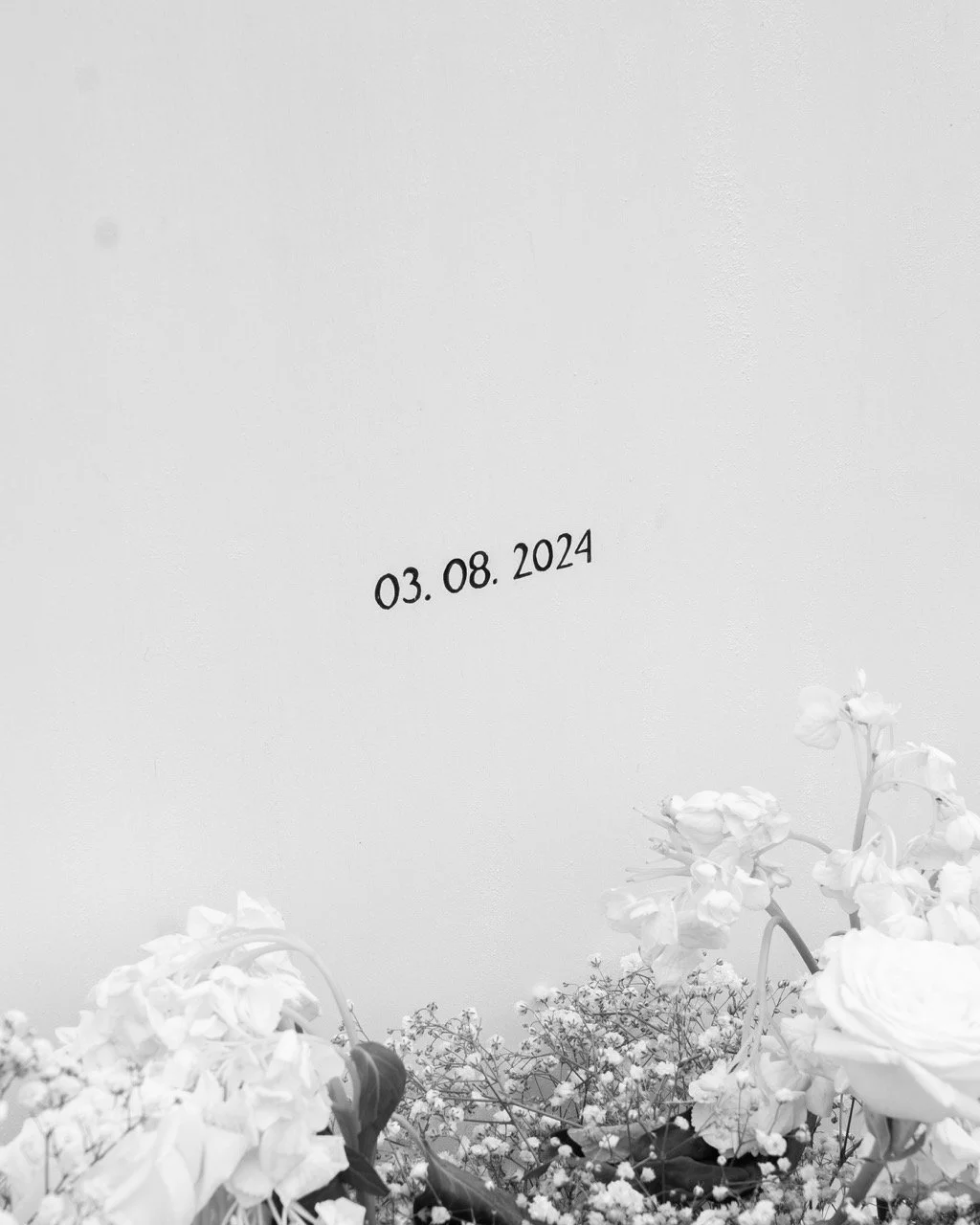 Black and white photo of a wall with a date '03.08.2024' and a floral arrangement at the bottom.