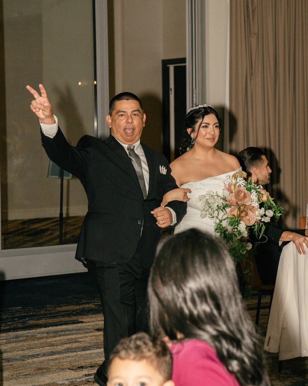 A joyous wedding celebration with a groom making a peace sign and a bride holding a bouquet of flowers, both dressed in formal wedding attire.