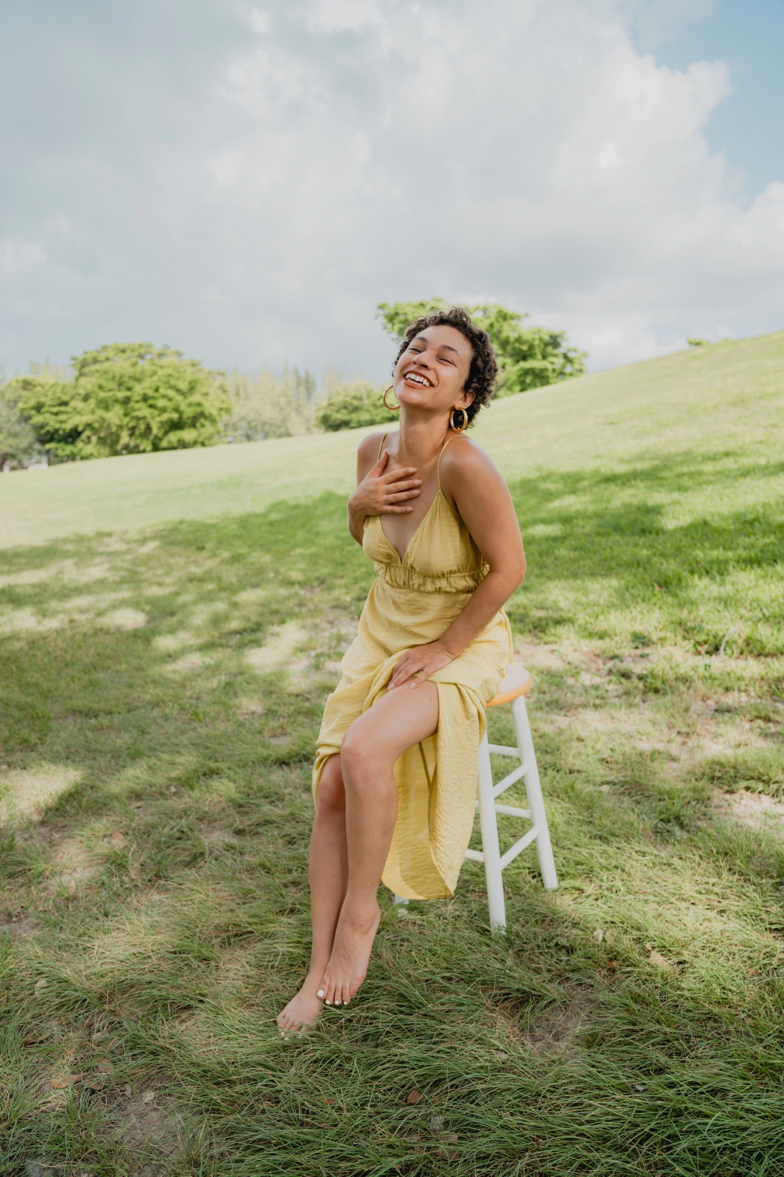 A woman in a yellow dress sitting on a white stool outdoors, smiling and laughing with her hand on her chest, on a grassy field under a partly cloudy sky.