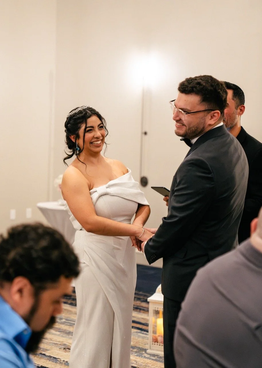 A bride and groom holding hands and smiling during their wedding ceremony in a formal setting, with a man in a black suit and glasses standing next to them and other guests visible in the foreground.