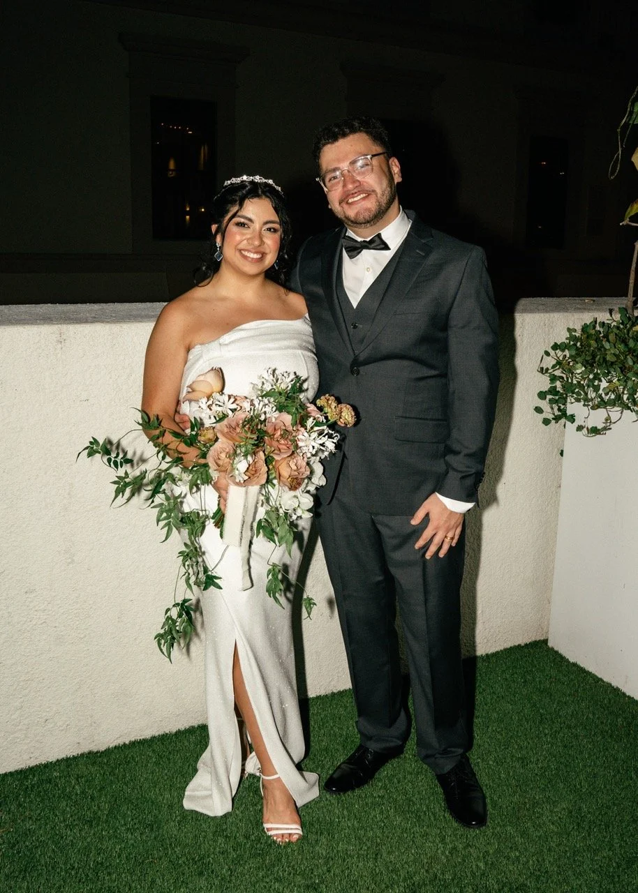 A bride and groom stand together at their wedding celebration, smiling for the camera. The bride wears a white strapless wedding gown, holds a bouquet of pink and white flowers, and has dark hair styled with a tiara. The groom wears a dark tuxedo wit