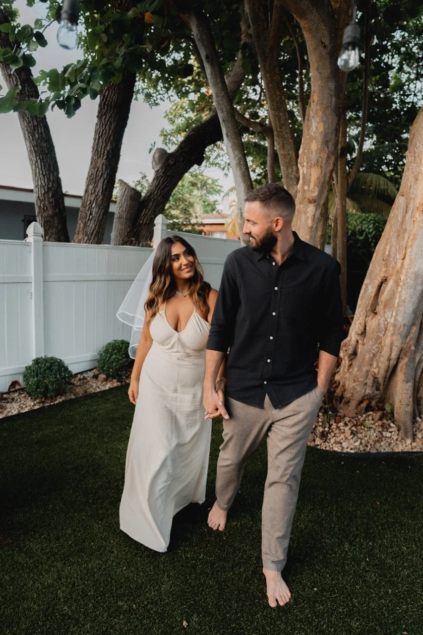 A woman in a white wedding dress and veil is holding hands with a man in a black shirt and gray pants, walking barefoot in a backyard with a white fence, green grass, and large trees.
