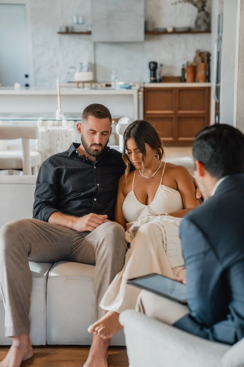 Couple holding hands during therapy session with therapist taking notes in a cozy, modern living room.