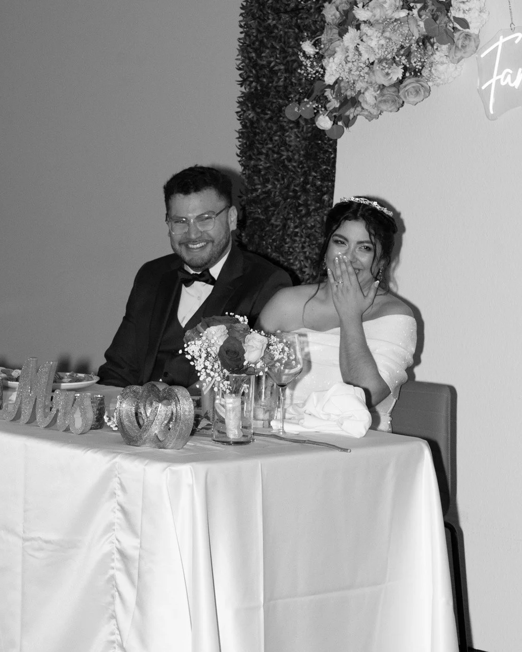 A black and white photo of a newlywed couple sitting at a wedding reception table, smiling and laughing. The man is in a tuxedo and the woman is in a wedding dress with a tiara. The table has floral arrangements, glasses, and decorative items.