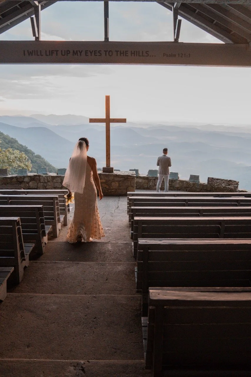 A bride stands inside a chapel, holding a bouquet, facing an altar with a large wooden cross and a mountain view outside. A man in a light suit stands near the altar facing away, overlooking the landscape. The chapel has wooden pews and a sign above 