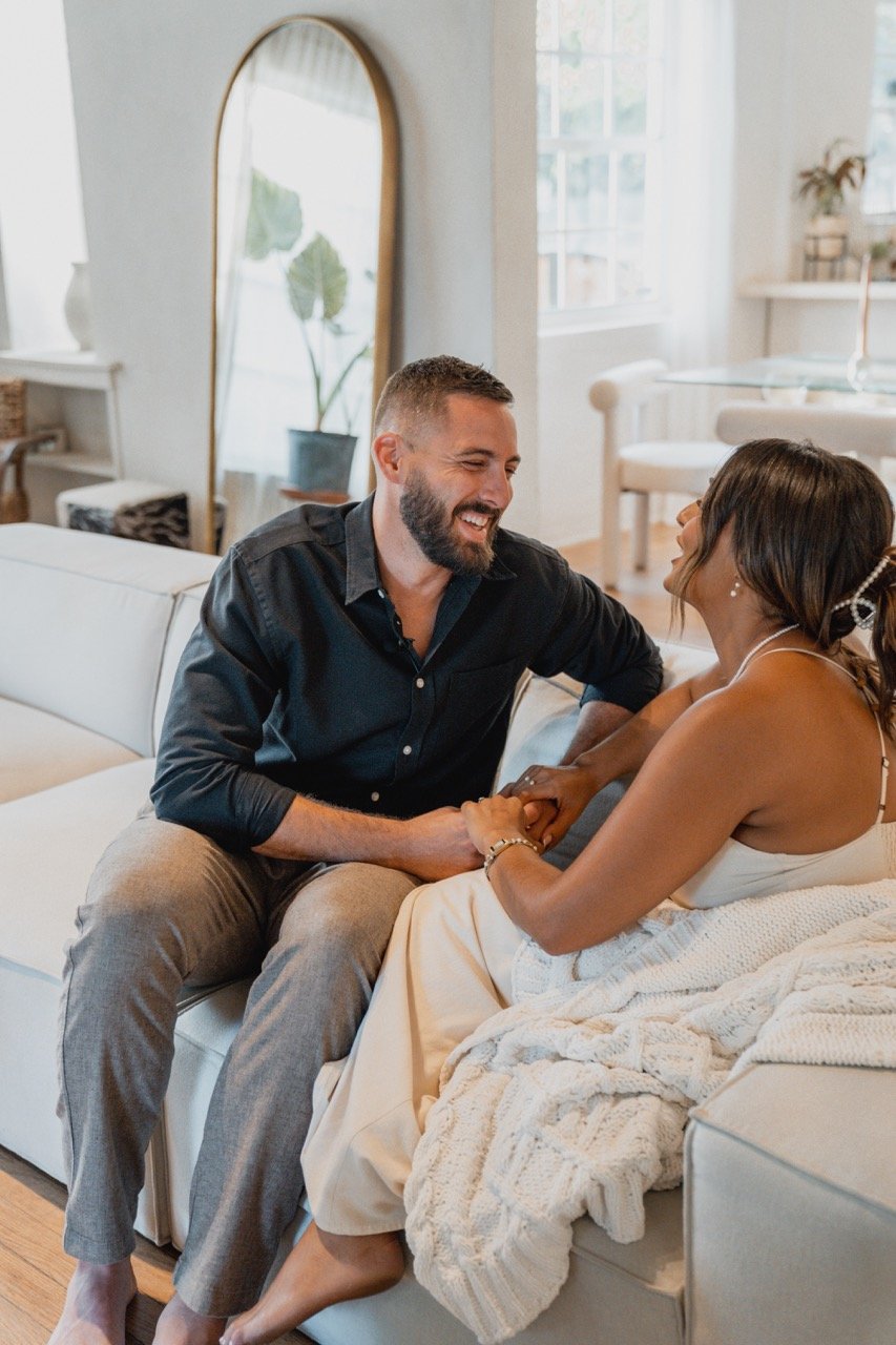 A couple sitting on a white sofa in a bright, cozy living room, holding hands and smiling at each other.
