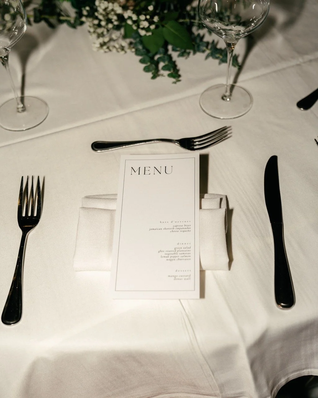 Elegant table setting with white tablecloth, neatly folded napkin, printed menu, two forks on the left, a knife and a fork on the right, and empty wine glasses at the top, with a floral centerpiece in the background.