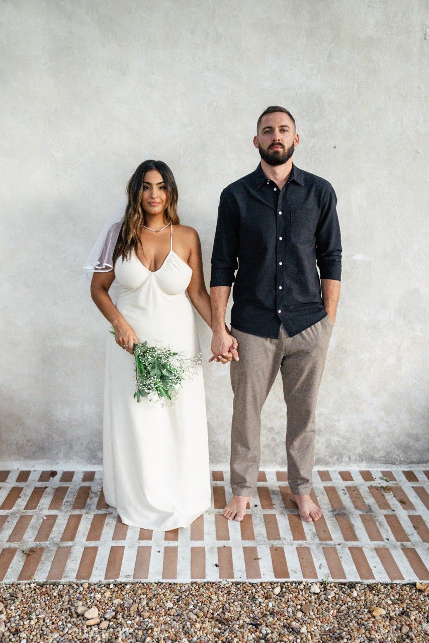 A couple standing hand-in-hand against a plain wall, with the woman wearing a white wedding dress and holding a bouquet, and the man dressed in a dark shirt and light pants, both barefoot.