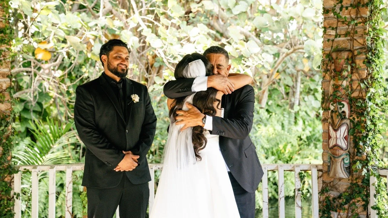 A wedding scene with three people: a bride in a white dress hugging an older man, possibly her father, and an officiant standing nearby. The background features lush green foliage and decorative tiles on a brick wall.