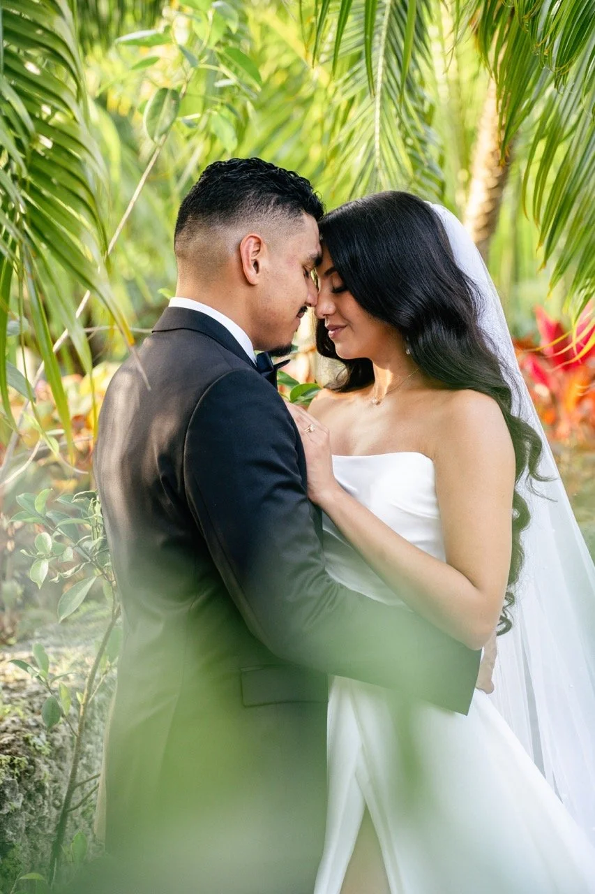 A bride and groom in a close, intimate pose outdoors surrounded by green tropical plants, with their foreheads touching and eyes closed.