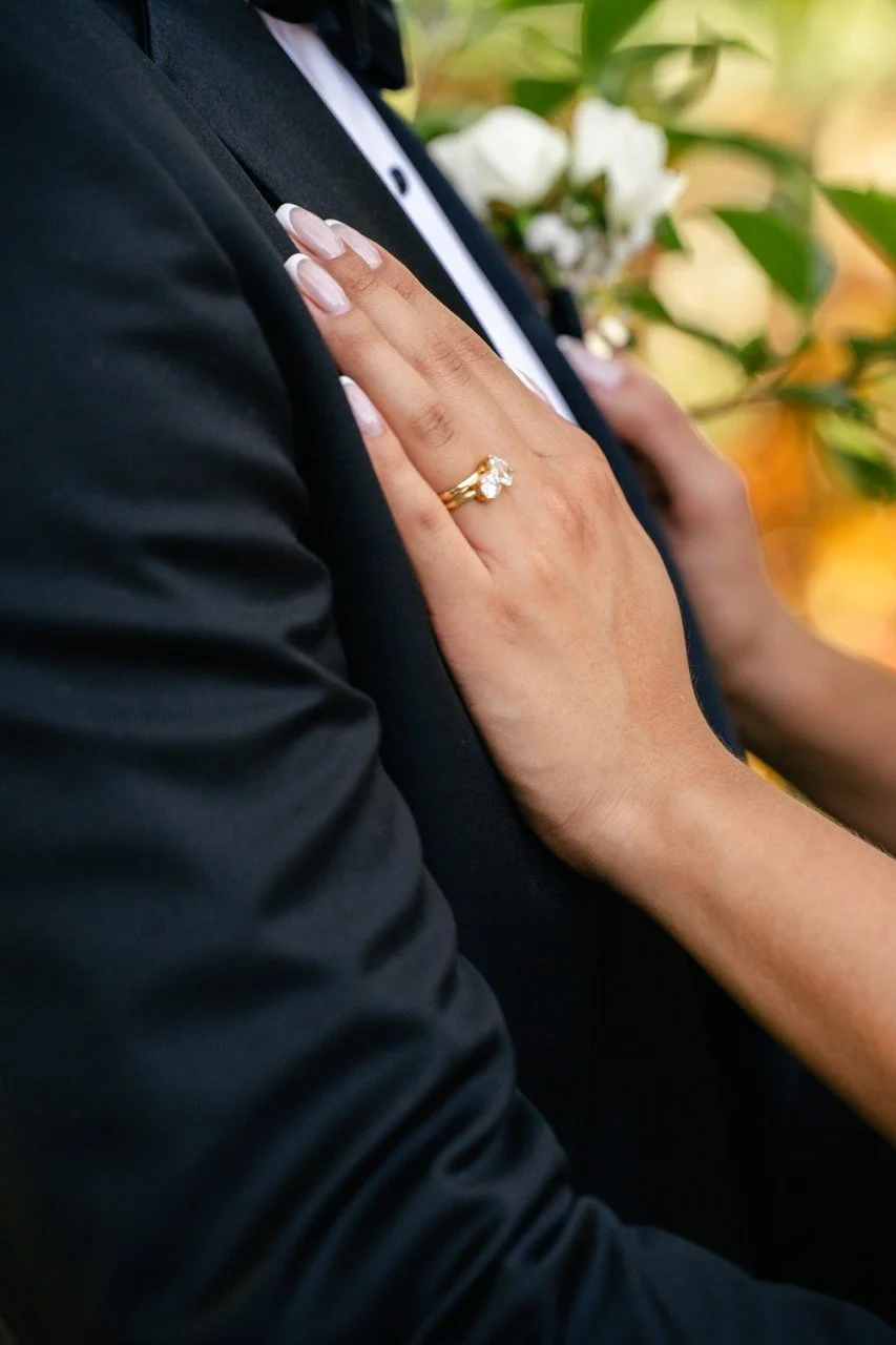 A close-up of a woman's hand resting on a man's chest, showing a wedding ring with a large central diamond. The man is wearing a black suit with a white shirt, and there are white flowers and green leaves in the blurred background.