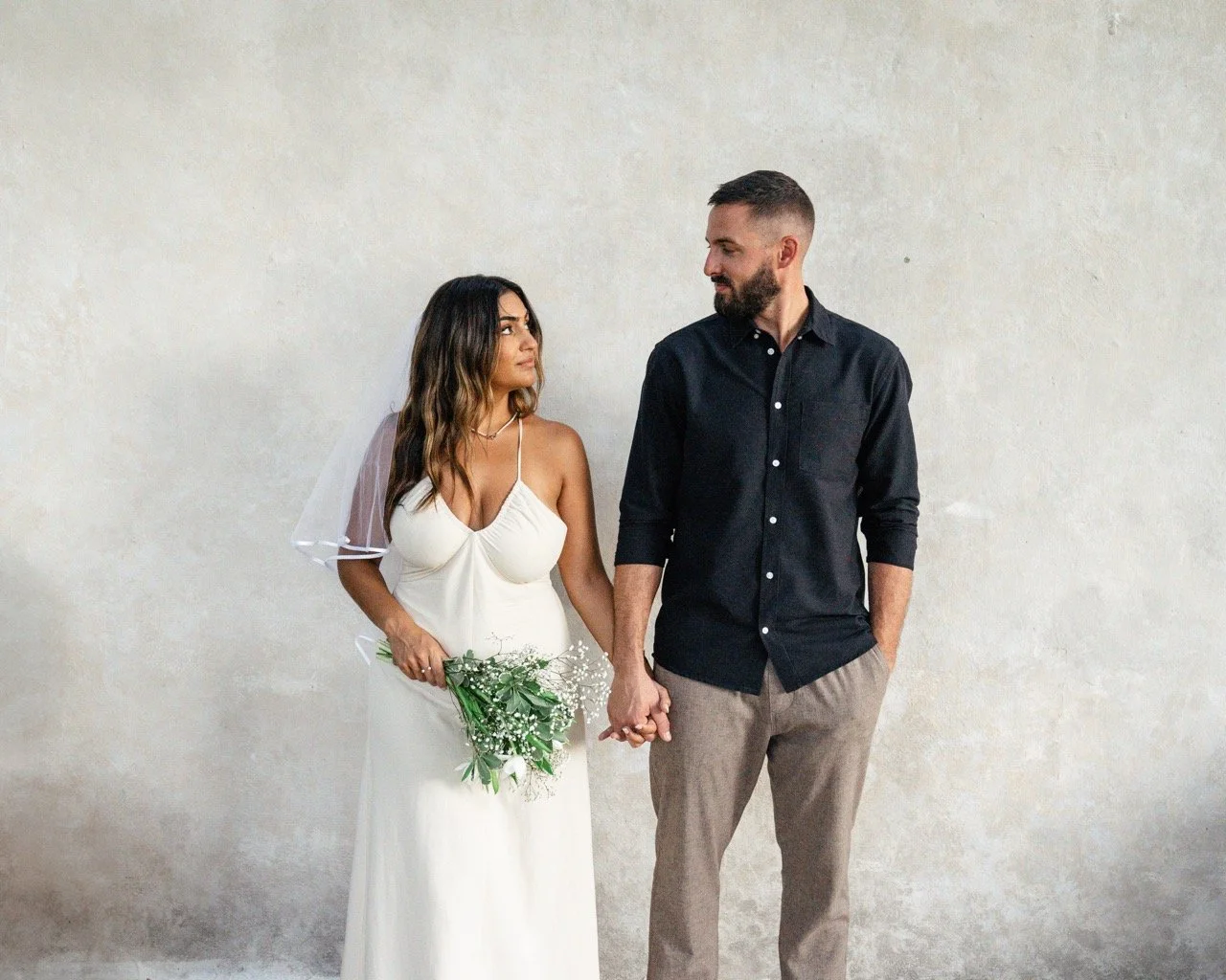 A woman in a white wedding dress holding a bouquet of flowers, standing next to a man in a black shirt and gray pants, holding hands and looking at each other against a plain, light-colored wall.