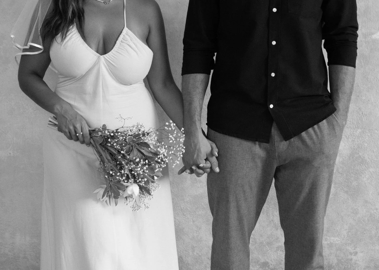 A couple holding hands, with the woman in a wedding dress holding a bouquet of flowers, and the man in a dark shirt and gray pants.