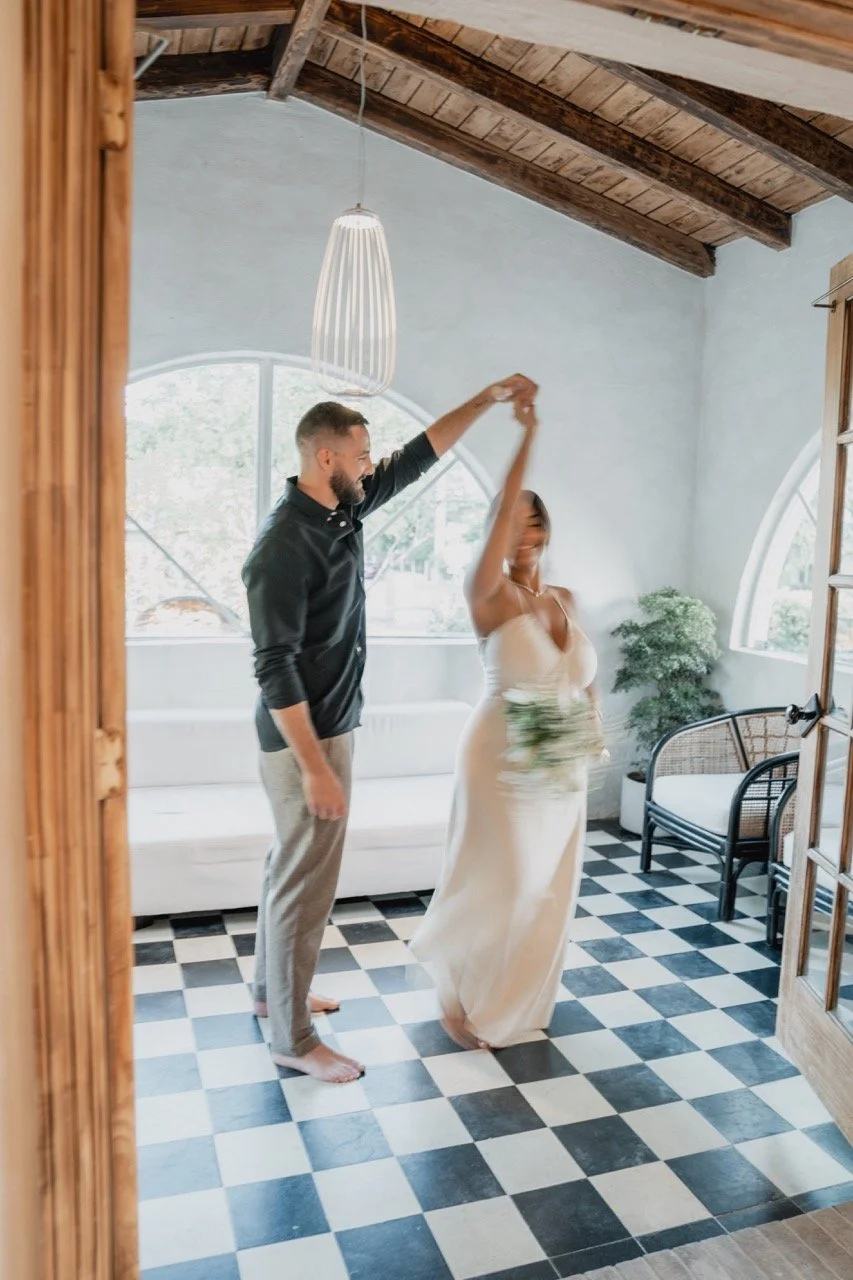 A couple dancing indoors, with the woman in a white wedding dress holding a bouquet and the man in a dark shirt and light pants, smiling in a bright room with large windows and checkered black-and-white floor tiles.