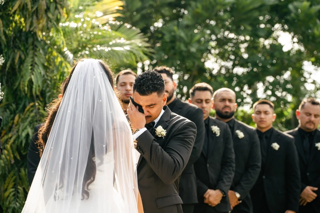 Man in a black suit crying during wedding ceremony, surrounded by groomsmen in black suits, outdoors with greenery