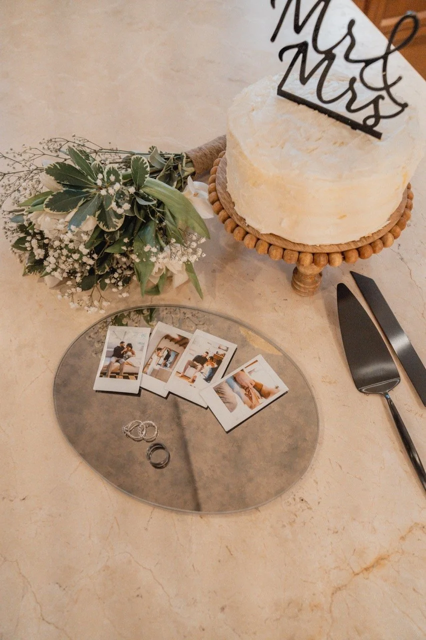 A wedding cake with a topper that reads 'Mr & Mrs,' a bouquet of flowers, Polaroid photos, two wedding rings, and a cake knife and server on a marble table.