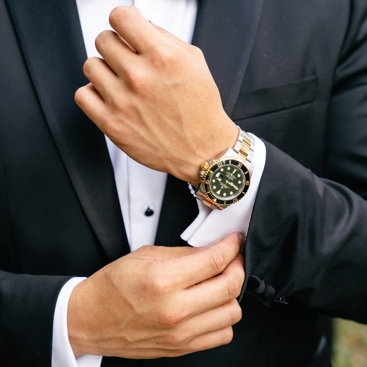 A man in a tuxedo adjusting his sleeve, wearing a luxury wristwatch with a black dial and gold accents.