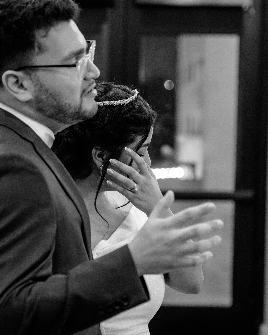 A couple, possibly at a wedding, with the man in a suit and glasses and the woman in a white dress and tiara, both with eyes closed, holding hands, appearing emotional.