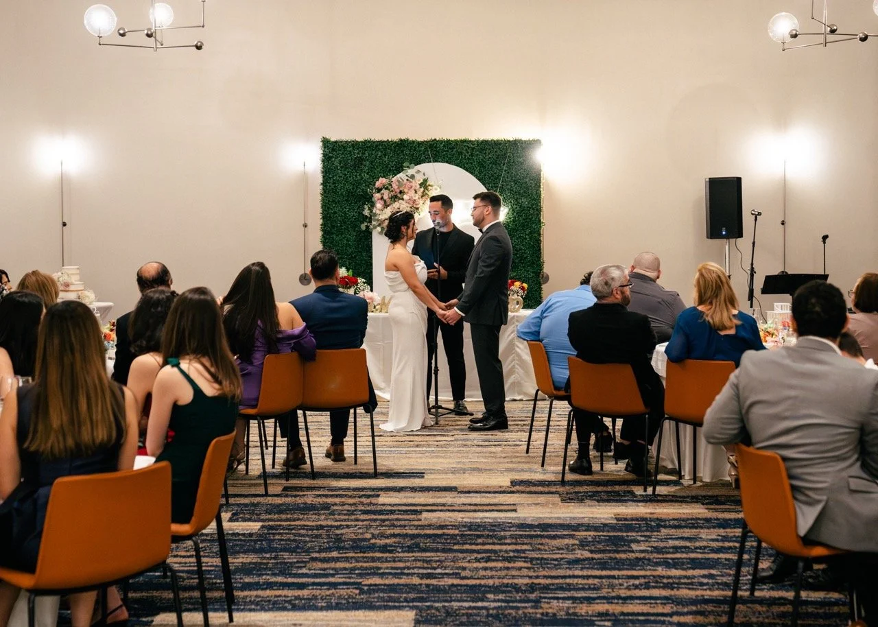 A wedding ceremony with a couple holding hands in front of an officiant, standing on a decorated stage, while guests seated at round tables watch. The room has beige walls, modern lighting, and a floral backdrop.