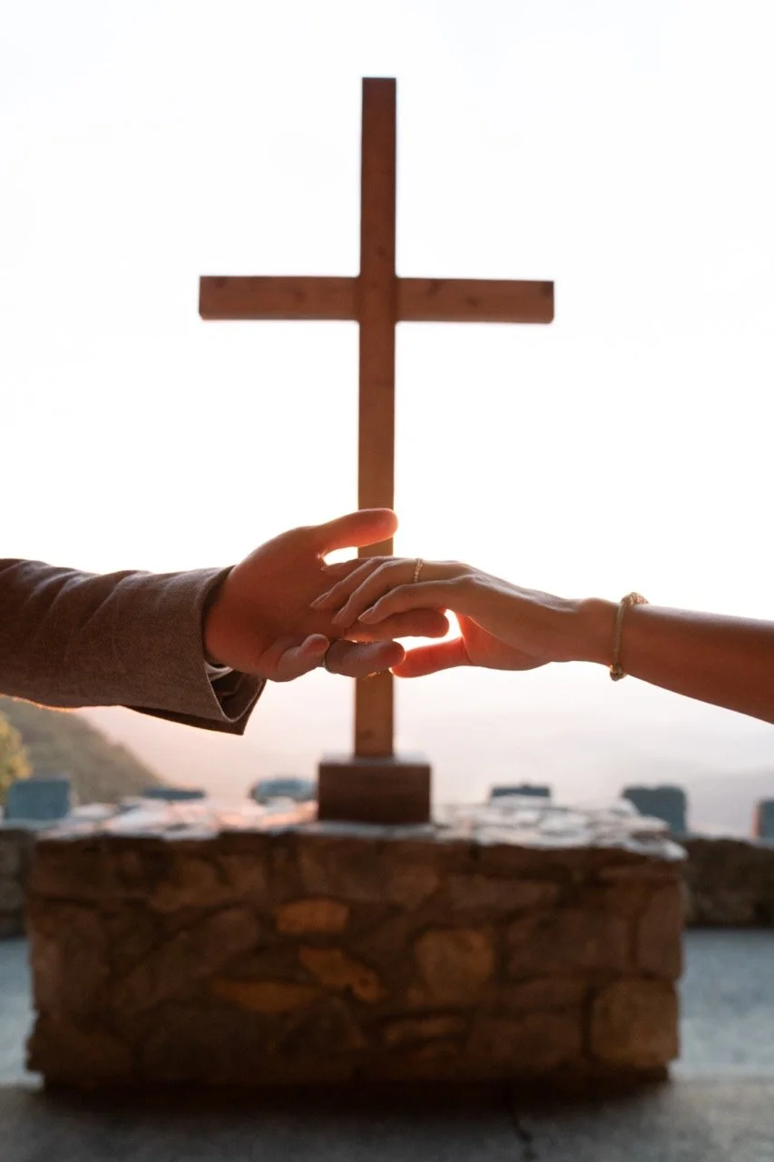 Two people holding hands in front of a wooden cross on a stone pedestal, with a body of water and boats in the background, at sunset.