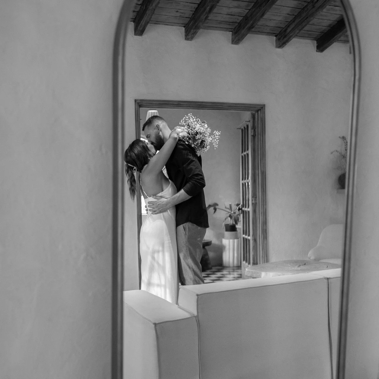 A black-and-white photo of a romantic moment reflected in a mirror, showing a couple embracing and kissing in a cozy room with wooden ceiling beams and potted plants.