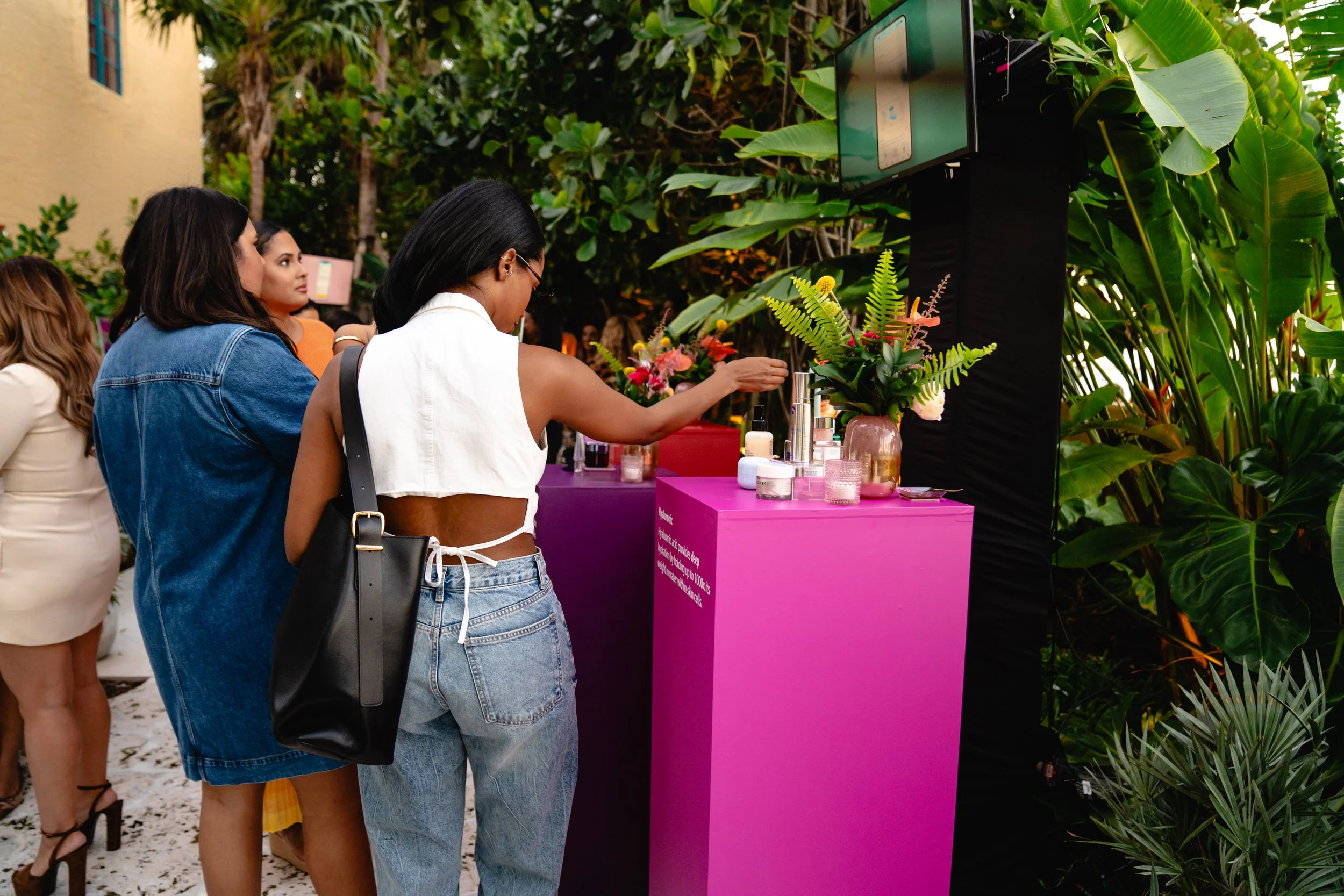 Women at a pink skincare or beauty product display table outdoors with green tropical plants in the background.