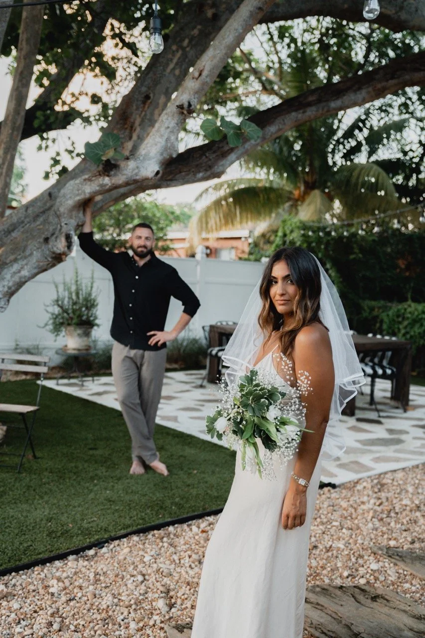 Bride in a white wedding dress holding a bouquet of flowers, standing outdoors in a garden, with a man casual dressed in black shirt and light pants in the background, under a large tree with hanging light bulbs.