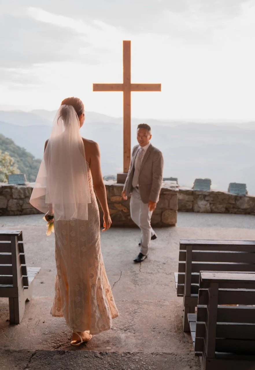 Bride and groom at an outdoor wedding ceremony with a large wooden cross and mountain scenery in the background.