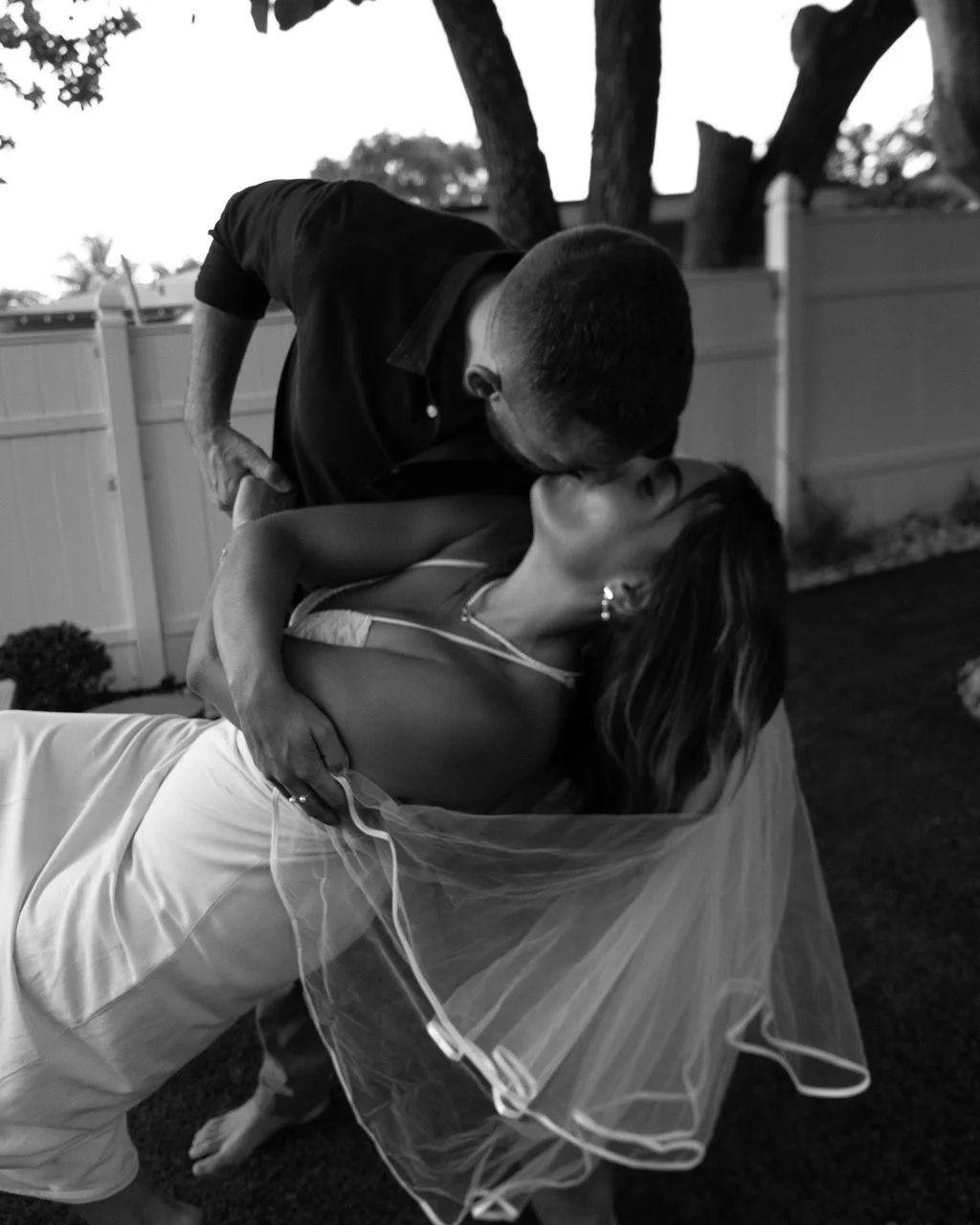 A man and woman sharing a kiss outdoors, with the man leaning over the woman who is lying back. The woman is dressed in a wedding gown with a veil, and the man is dressed in dark clothing. They are near a white fence and trees.