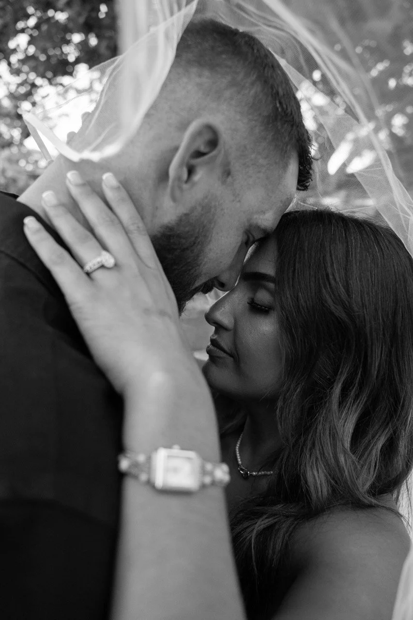 A black-and-white close-up photo of a couple embracing each other, their foreheads touching, with a veil draped over them. The woman has long dark hair and wears a necklace and a watch, and the man has short hair and a beard.