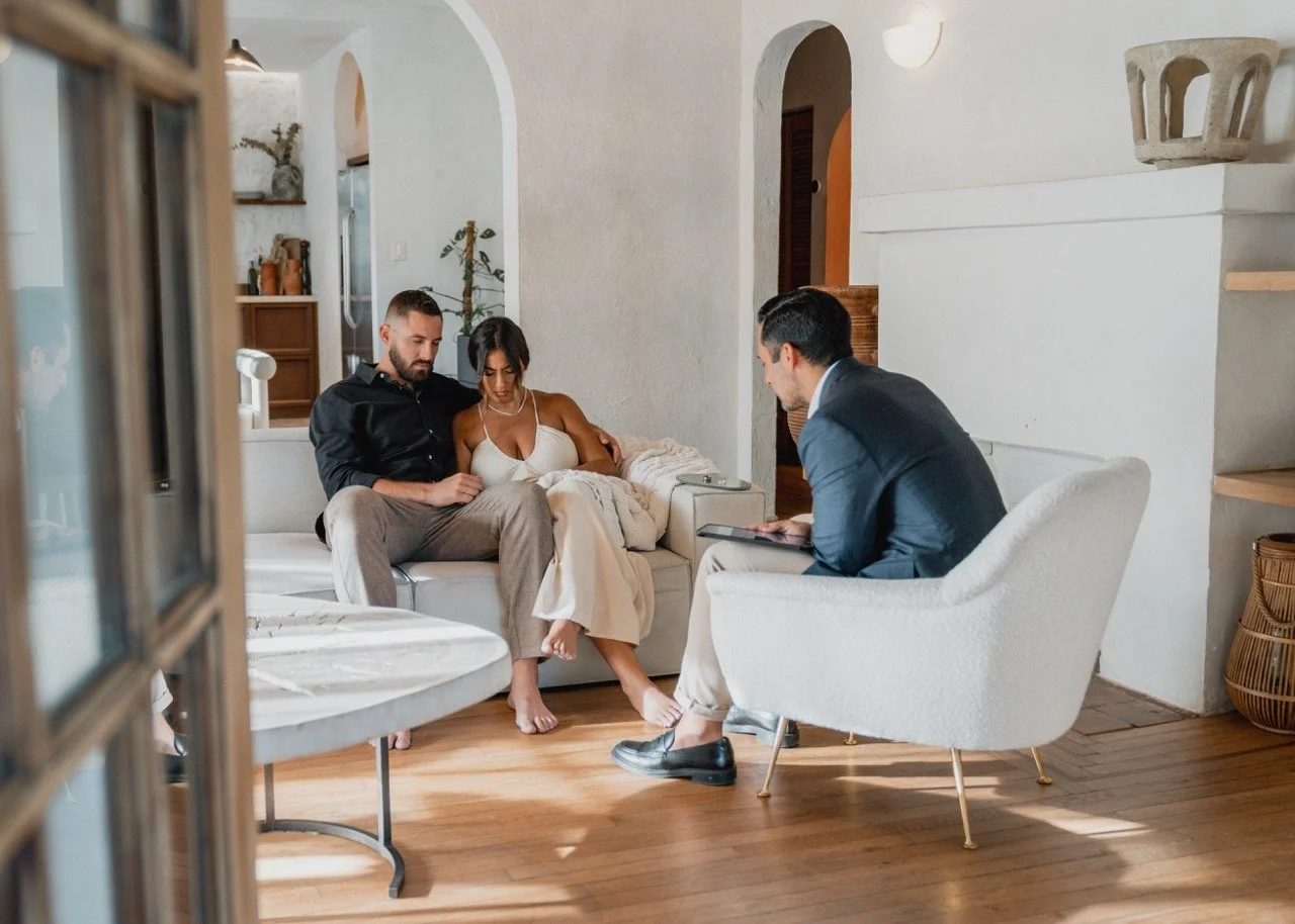 A couple sits on a white sofa, talking with a therapist who is sitting in a white armchair, in a cozy living room with wooden floors and white walls.