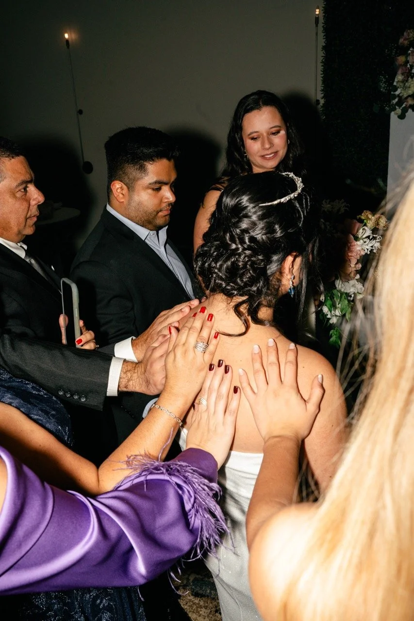 People gathered around a bride, touching her shoulder and back in a celebratory moment, likely at a wedding reception.