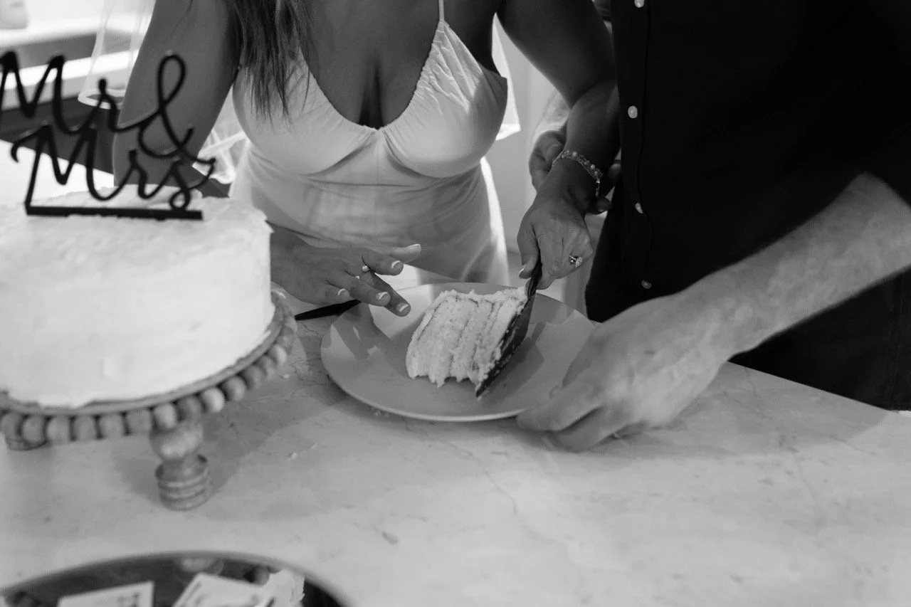 A couple is cutting a slice of wedding cake together at a celebration. The cake has a 'Mr & Mrs' topper, and the scene is in black and white.
