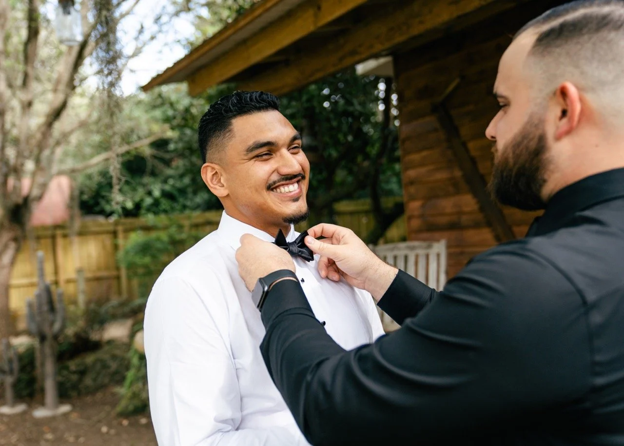 Two men, one in a white dress shirt and the other in a black shirt, outdoors in a backyard. The man in the black shirt is adjusting the bow tie of the man in the white shirt, who is smiling.